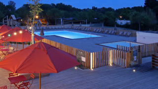 Vista nocturna de Camping d'Angers - Lac de Maine con piscina iluminada y terraza con sombrillas rojas.