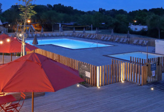 Evening view of Camping d'Angers - Lac de Maine with illuminated pool and terrace featuring red umbrellas.
