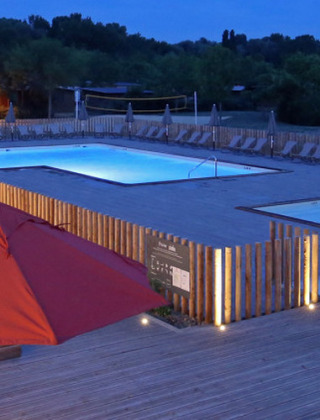 Vista nocturna de Camping d'Angers - Lac de Maine con piscina iluminada y terraza con sombrillas rojas.