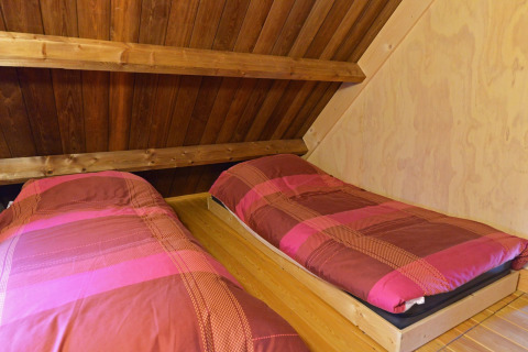 To enkeltsenge med rød- og pinkfarvet sengetøj under et skråt trætag i et træhus på Camping de Koeksebelt.