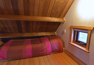 Acogedor dormitorio con techo inclinado de madera, cama individual y pequeña ventana en una casa del árbol.