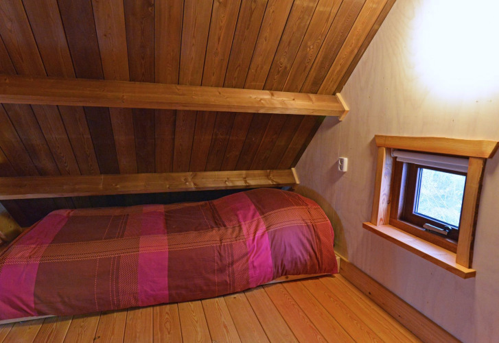 Knusse slaapkamer met schuin houten plafond, eenpersoonsbed en klein raam in een boomhut in Nederland.