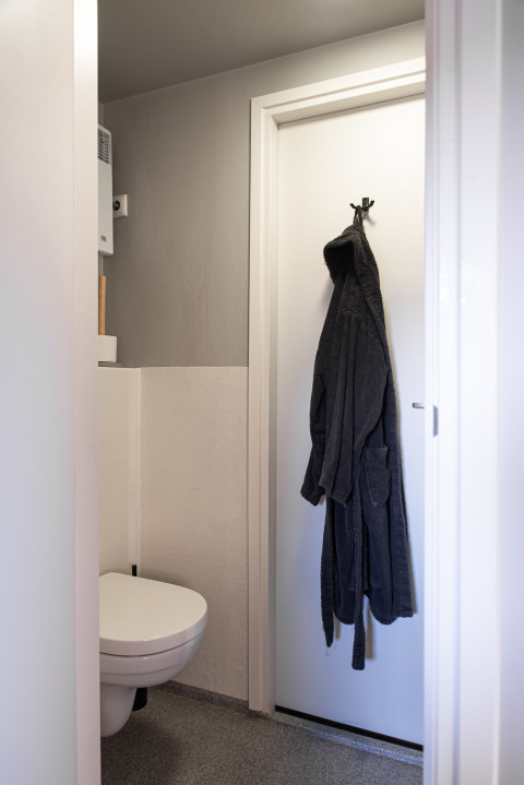 Lille badeværelse med væghængt toilet og mørk badekåbe på en hvid dør i Wellness Lodge, Ackerlodges Ruinerwold BV.