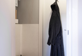 Lille badeværelse med væghængt toilet og mørk badekåbe på en hvid dør i Wellness Lodge, Ackerlodges Ruinerwold BV.