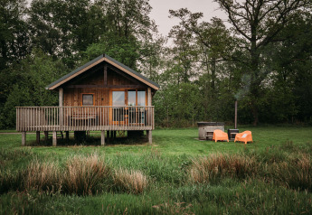 Wellness lodge med træhytte, veranda, orange stole og udendørs badekar omgivet af grøn natur.
