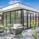 Moderne Forest villa-lodge med store glasvinduer, udendørs siddeområde og grøn have under blå himmel.