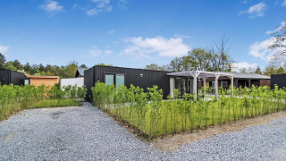 Eco Lodge moderna con fachada negra, camino de grava, setos verdes y cielo azul con algunas nubes.