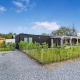 Moderne Eco Lodge med sort facade, grøn beplantning og grusvej under en blå himmel med spredte skyer.