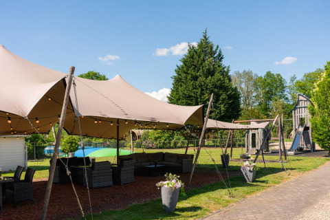 Outdoor lounge area with canopy, seating, and playground in a green holiday park setting in Overijssel, Netherlands.