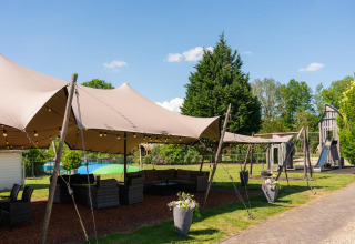Espace salon extérieur couvert, mobilier et aire de jeux dans un parc de vacances verdoyant à Overijssel, Pays-Bas.