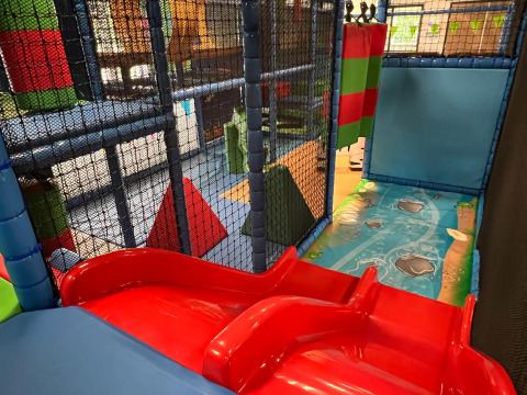 Parque de juegos interior con tobogán rojo, bloques acolchados y redes de seguridad en un parque vacacional.