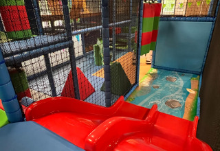 Parque de juegos interior con tobogán rojo, bloques acolchados y redes de seguridad en un parque vacacional.