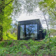 Nokken Forest Cabin, et sort tiny house med store vinduer i Wilsumer Berge, Tyskland omgivet af grøn skov.