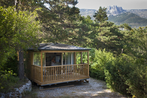 Acogedora cabaña de madera en Huttopia Gorges du Verdon, rodeada de vegetación y con montañas al fondo.