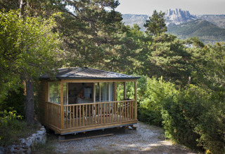 A cozy wooden cabin nestled in greenery at Huttopia Gorges du Verdon holiday park, with mountains in the background.