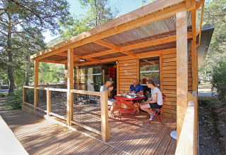 Feriehytte i træ med overdækket terrasse, hvor folk nyder udendørs måltid omgivet af naturen.