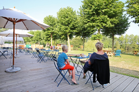 Zona de cafetería al aire libre con mesas, sombrillas y personas en Huttopia Baie du Mont St Michel, Bretaña, Francia.