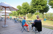 Zona de cafetería al aire libre con mesas, sombrillas y personas en Huttopia Baie du Mont St Michel, Bretaña, Francia.