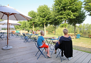 Zona de cafetería al aire libre con mesas, sombrillas y personas en Huttopia Baie du Mont St Michel, Bretaña, Francia.