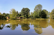 Casette di legno vicino a un lago con riflessi di alberi e cielo nell’acqua a Huttopia Baie du Mont St Michel, Bretagna, Francia.
