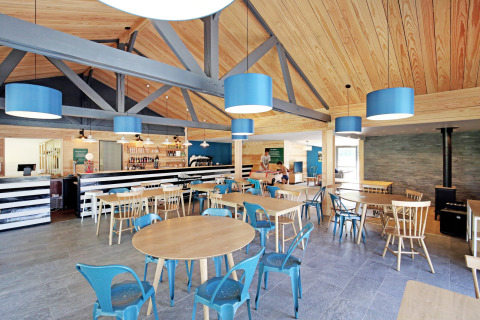 Café moderne avec plafond en bois, lampes bleues et chaises variées à Huttopia Baie du Mont St Michel, Bretagne.