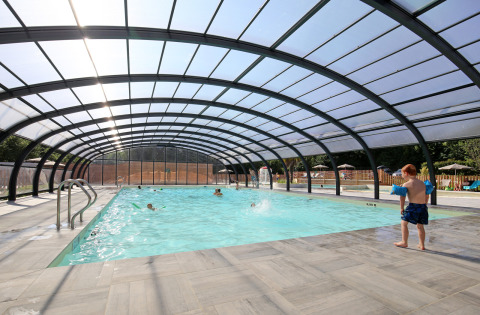 Indoor pool with glass roof, children swimming and a boy preparing to enter the water at Huttopia Baie du Mont St Michel.