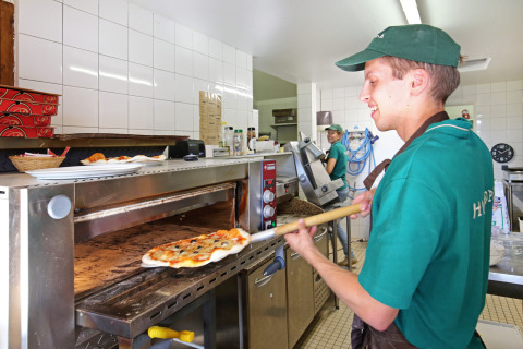 Un pizzero con uniforme verde saca una pizza recién hecha del horno en Huttopia Baie du Mont St Michel.
