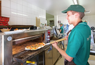 Un pizzero con uniforme verde saca una pizza recién hecha del horno en Huttopia Baie du Mont St Michel.
