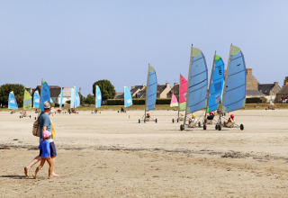 En far og et barn går på stranden ved Huttopia Baie du Mont St Michel i Bretagne, Frankrig, med sejlvogne i baggrunden.