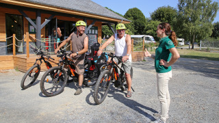 Dos ciclistas con cascos conversan con una empleada frente a una cabaña en Huttopia Baie du Mont St Michel.