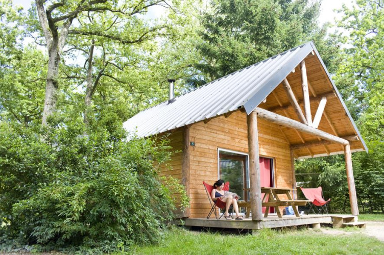 Wooden cabin surrounded by lush greenery and trees with a person relaxing outside at Huttopia Rambouillet, France.