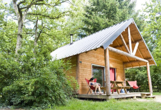 Gemütliche Holzhütte im Grünen mit einer Person, die entspannt davor sitzt, aufgenommen im Huttopia Rambouillet.