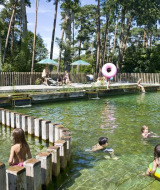 Niños nadan y juegan en una piscina natural rodeada de árboles en Huttopia Rambouillet, Île de France.