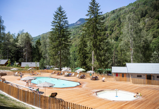 Udendørs poolområde med liggestole og parasoller i Huttopia Bozel en Vanoise, omgivet af grønne bjerge.