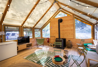 Vue intérieure d'une tente glamping chaleureuse avec sol en bois, coin salon, guirlandes lumineuses et fenêtres.