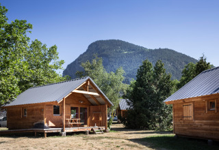 Wooden cabins and picnic table with mountain and trees at Huttopia Bozel en Vanoise, Auvergne-Rhône-Alpes, France.