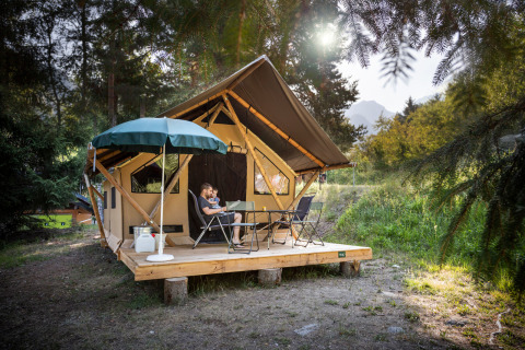 Family relaxes on wooden deck in front of a Canadienne-style safari tent surrounded by forest and sun.