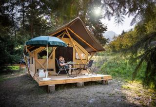 Familia relajándose en terraza de madera frente a una tienda safari Canadienne rodeada de naturaleza y sol.