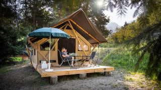 Familia relajándose en terraza de madera frente a una tienda safari Canadienne rodeada de naturaleza y sol.
