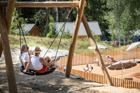 Two people relax on a large swing near a holiday park with wooden cabins and sunbathing decks in France.