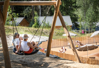 Twee personen genieten op een schommel bij houten huisjes en terras in een vakantiedomein in Frankrijk.