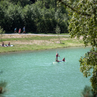 Personas remando en lago cerca de Bozel, Francia, con árboles verdes y caminantes al fondo.
