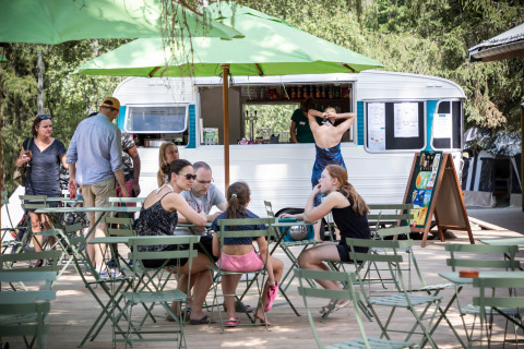 Café al aire libre con mesas verdes, personas y una caravana de comida en un parque vacacional francés.