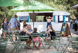 Udendørs caféområde ved en foodtruck med folk, der slapper af under grønne parasoller i en feriepark.