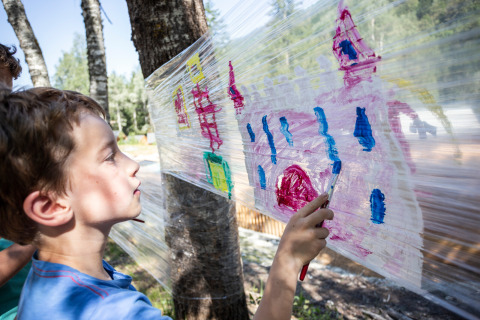 Boy painting on plastic wrap stretched between trees outdoors at Huttopia Bozel en Vanoise holiday park, France.
