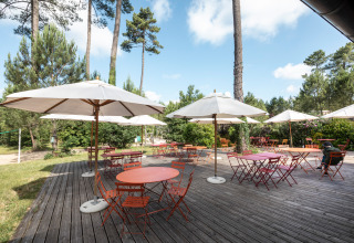 Sonnige Terrasse mit roten Tischen, Stühlen und weißen Sonnenschirmen im Ferienpark Huttopia Landes Sud.