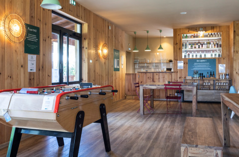 Acogedora sala común en Huttopia Landes Sud con futbolín, decoración de madera y barra de bebidas.