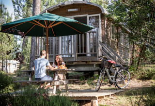 Pareja desayunando en una mesa de picnic frente a una cabaña Roulotte en Huttopia Landes Sud, Francia, rodeados de bosque.