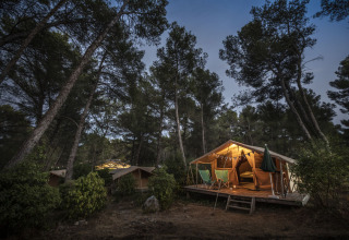 Vista nocturna de la tienda safari Bungalow Classic en Huttopia Fontvieille, Francia, rodeada de pinos altos.