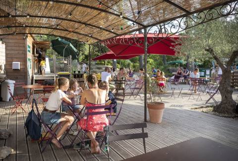 Gezellige buitenterras met mensen aan tafels onder parasols bij Huttopia Fontvieille, Frankrijk.
